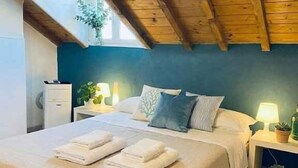 Un dormitori amb sostre de fusta, un llit amb roba blanca, una tauleta de nit amb una làmpada i una finestra amb una planta.