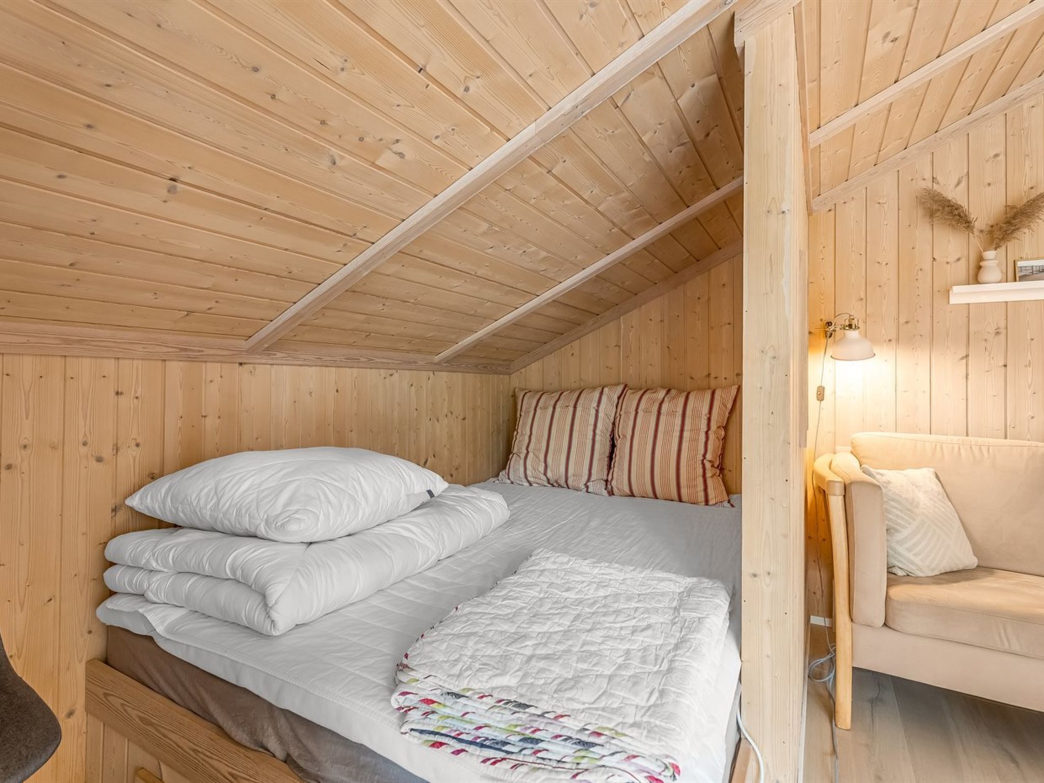 A wooden cabin bedroom with a bed, pillows, a quilt, a sofa, and a wall-mounted lamp.