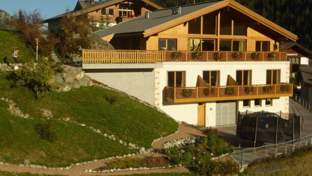 Een houten chalet met twee verdiepingen, voorzien van balkons, een pad van stenen en een bergachtige achtergrond.