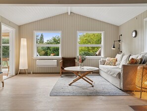 Ein Zimmer mit schrägen Wänden, zwei Fenstern mit Blick ins Grüne, einem weißen Sofa mit gemusterten Kissen, einem runden Couchtisch und einem Beistelltisch mit Lampe.