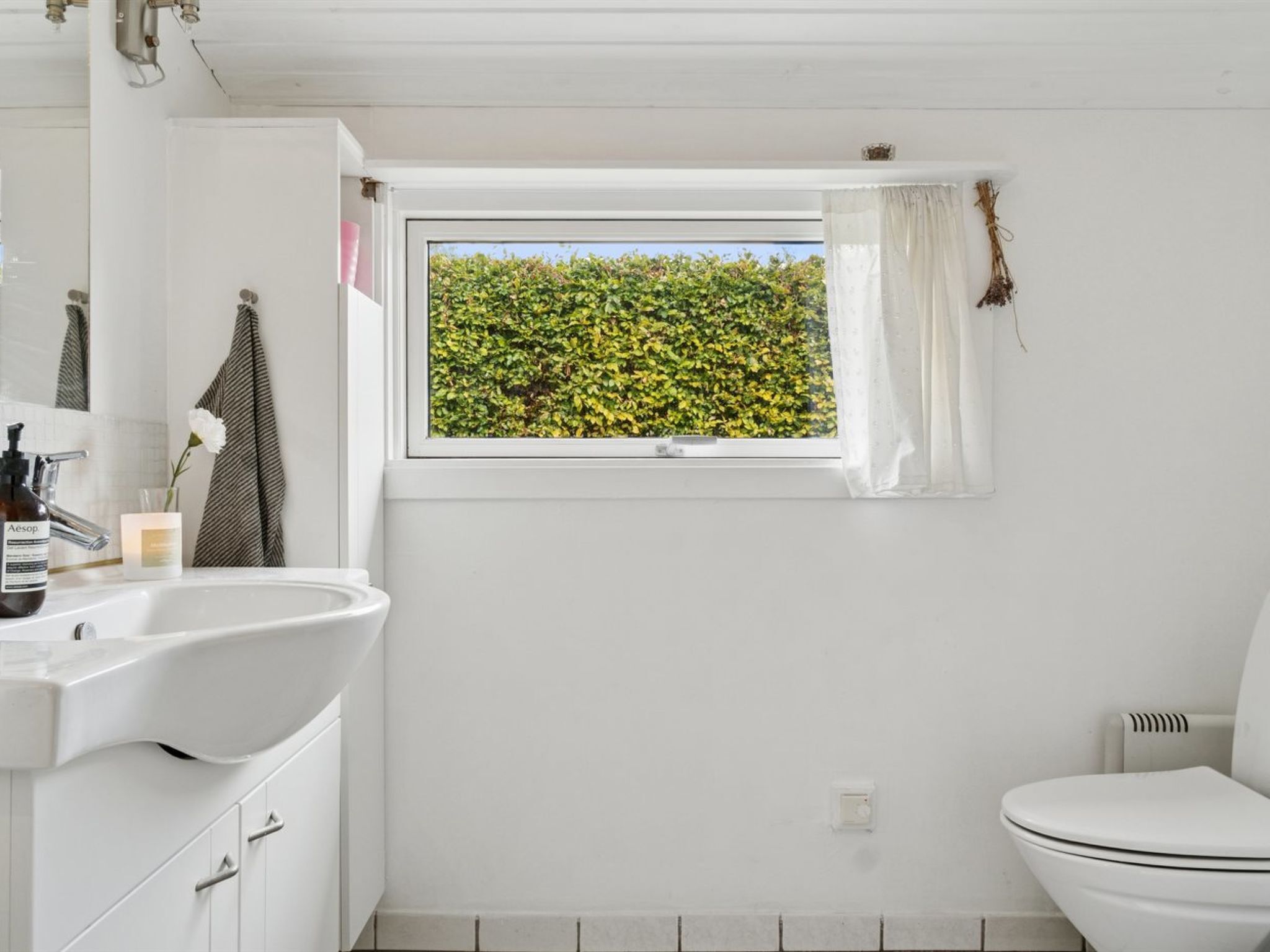 Ein Badezimmer mit einem weißen Waschbecken, einer Toilette, einem Fenster mit Blick auf Grünflächen und einem Handtuch an der Wand.