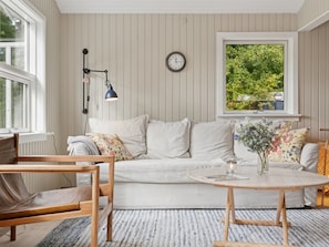 Ein Zimmer mit einer weißen Couch, einem Holzstuhl, einem runden Tisch, einer Wanduhr und einem Fenster mit Blick auf Grünflächen.