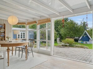 Ein Wintergarten mit Holztisch und -stühlen, großen Fenstern und Blick auf einen Garten mit einem blauen Gebäude.