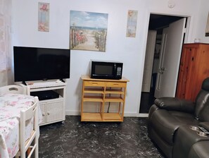 A living room with a television, a microwave on a wooden stand, a leather sofa, a wicker table with a floral tablecloth, and a painting on the wall. - Cozy 1-bedroom Cottage furnished with all utilities included (Port Orange)