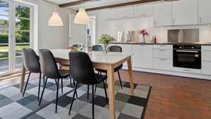 Moderni keittiö, jossa on valkoiset kaapit, ruostumattomasta teräksestä valmistettu uuni sekä ruokailutila, jossa on pöytä ja tuolit.
