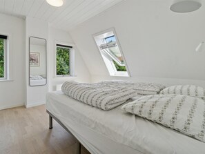 Ein Schlafzimmer mit schrägen Wänden, einem Bett mit gemusterten Textilien, einem großen Spiegel und einem Fenster mit Blick auf Grünfläche.
