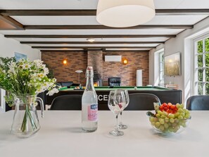 Ein Billardtisch mit grüner Spielvliesbahn, eine Glasflasche, zwei Weingläser und eine Obstschale auf einem weißen Tisch.