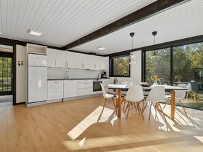 Eine moderne Küche mit weißen Schränken, ein Essbereich mit einem Holztisch und Stühlen sowie große Fenster mit Blick ins Freie.