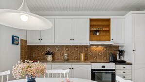 A kitchen with white cabinets, a wooden shelf, a coffee maker, and a vase with flowers on the table. - "Hening" - 150m from the sea by Interhome (Allinge)