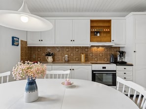 Una cocina con gabinetes blancos, un estante de madera, una cafetera y un florero con flores sobre la mesa.