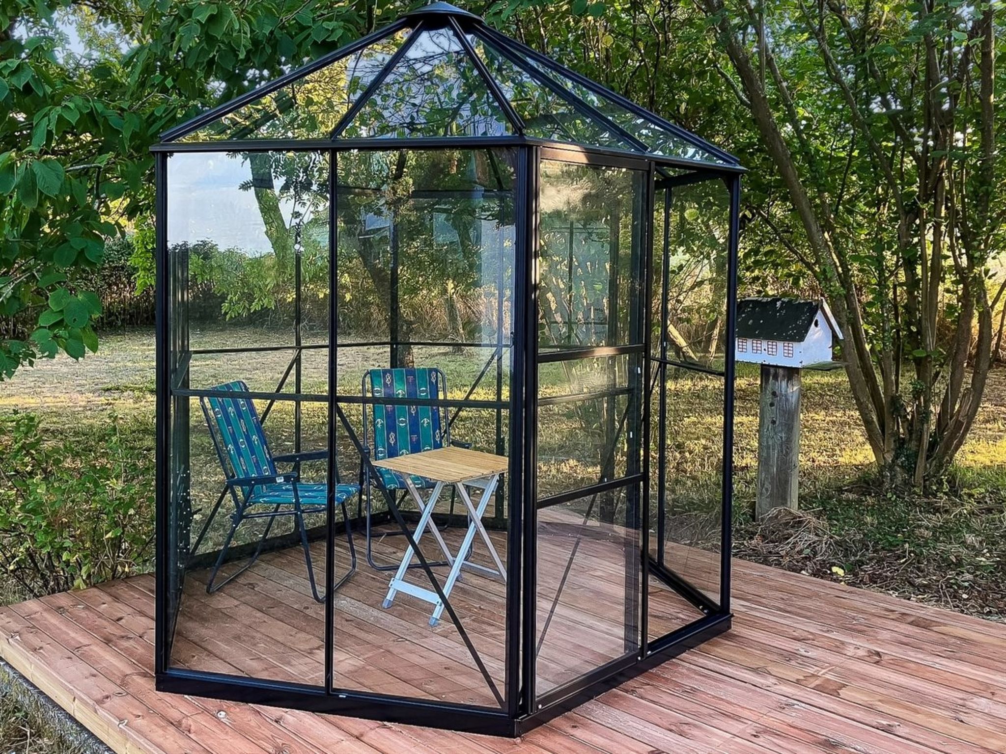A glass-enclosed gazebo with a wooden deck, two blue chairs, and a small table.