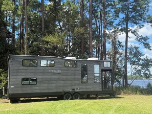 A mobile home with a kitchenette, a sleeping area, and a bathroom. It is parked on grass with trees and a lake in the background.