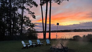 A sunset view with a lake, trees, and three Adirondack chairs facing the water.