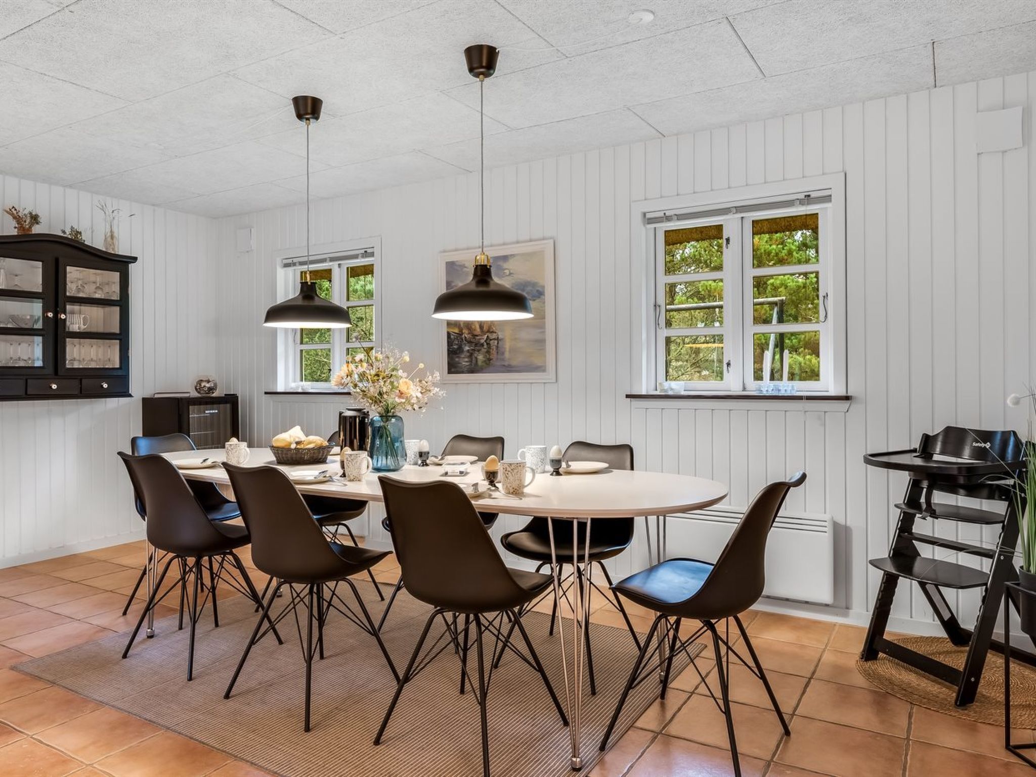 A dining area with a round table, six chairs, pendant lights, a cabinet, and windows with greenery outside.
