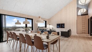 Un espace repas moderne avec une grande table en bois, plusieurs chaises, des luminaires suspendus et un téléviseur fixé au mur.
