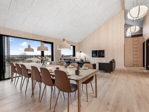 Ein moderner Essbereich mit einem großen Holztisch, mehreren Stühlen, Pendelleuchten und einem Fernseher an der Wand.