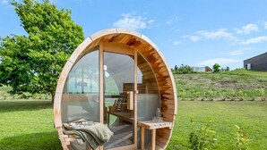 Une structure circulaire en bois, dotée de parois en verre, comprenant un banc et une table, installée sur une terrasse en bois.