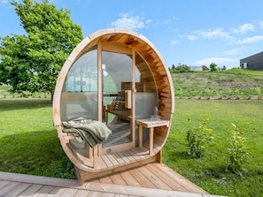Eine hölzerne, runde Konstruktion mit Glaswänden, ausgestattet mit einer Bank und einem Tisch, auf einer Holzterrasse platziert.