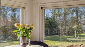 Ett rum med ett runt bord, en stol, en vas med blommor och utsikt över en trädgård genom stora fönster.