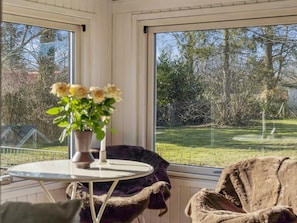 Ein Zimmer mit einem runden Tisch, einem Stuhl, einer Vase mit Blumen und Blick auf einen Garten durch große Fenster.