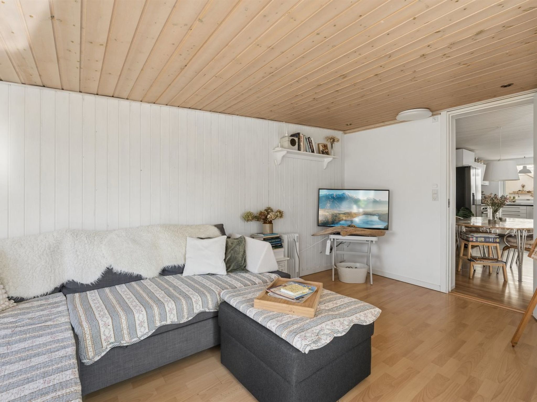 Un salon moderne avec un plafond en bois, un canapé d’angle gris, un téléviseur à écran plat et un coin repas visible à travers une porte ouverte.