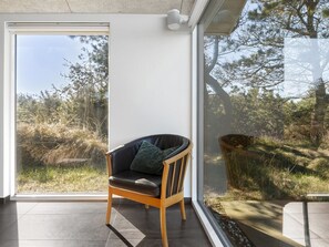 Ein Zimmer mit einem großen Glasfenster, das zu einer natürlichen Landschaft führt, ein Holzsessel mit Kissen und eine Beton Decke.