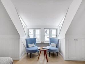 Een kamer met twee blauwe stoelen, een klein rond tafeltje met een boek, en een raam met uitzicht op gebouwen.