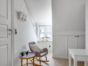 Una habitación con una puerta blanca, una pequeña mesa redonda con una silla, una ventana con vistas y una pared blanca con un timbre.