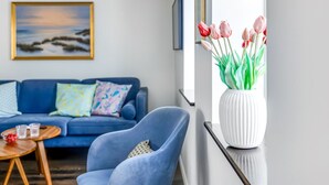 En stue med en blå sofa, en blå lænestol, en hvid vase med blomster og et maleri på væggen.