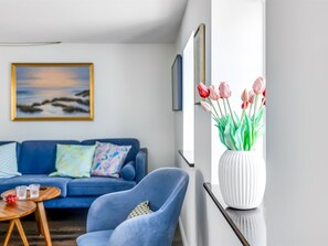 Una sala de estar con un sofá azul, un sillón azul, un jarrón blanco con flores y un cuadro en la pared.