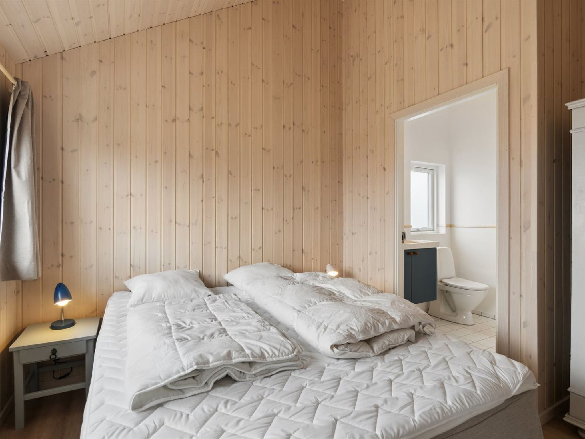 Una habitació amb un mur revestit de fusta, un llit amb roba de llit blanca, una làmpada de nit, un mirall i un vàter.