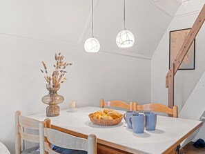 Un coin repas comprenant une table, des chaises, un vase avec des plantes séchées, un panier de pain et deux lampes suspendues.