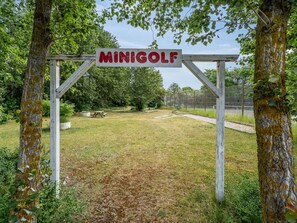 Une arche en bois sur laquelle est inscrit le mot « MINIGOLF », qui mène à un espace herbeux avec une table de pique-nique et des arbres.