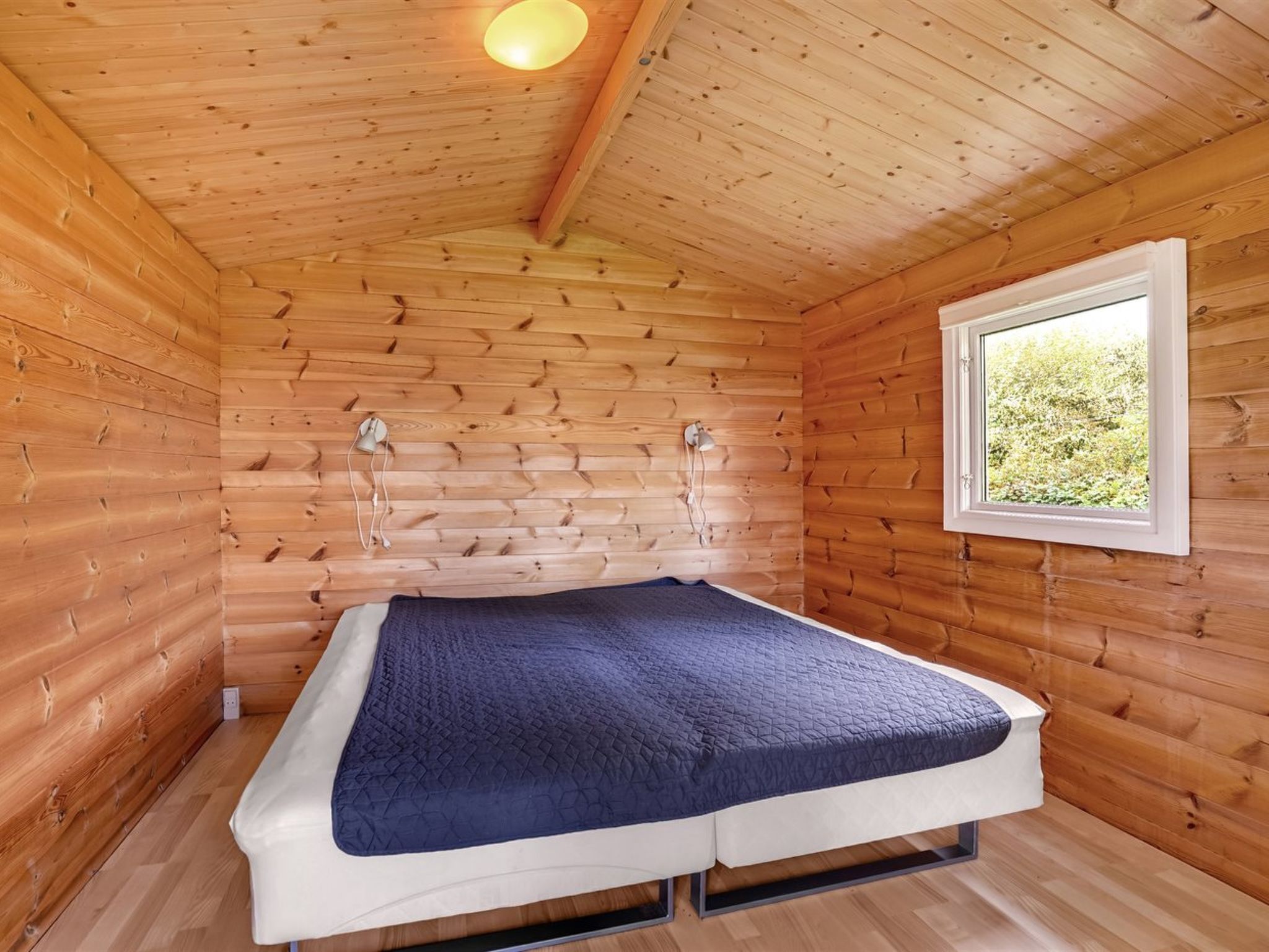 A small wooden cabin room with a bed, a window, and a light fixture.