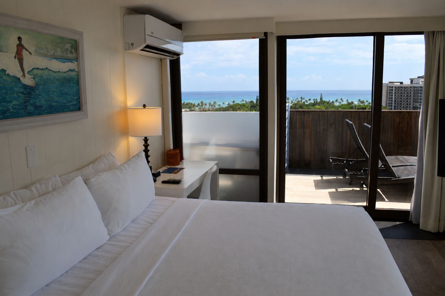 A hotel room with a large bed, a painting of a surfer, an air conditioning unit, a desk with a lamp, and a view of the ocean through a sliding door.