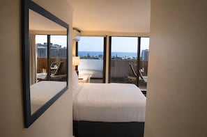 Une chambre d’hôtel avec un lit, un bureau et une chaise. Une grande fenêtre offre une vue sur les immeubles et l’océan.
