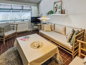 Ein gemütliches Zimmer mit einer Couch, einem Tisch mit Unterlage und Gläsern, einem Fernseher, einer Pflanze und einem Poster an der Wand.