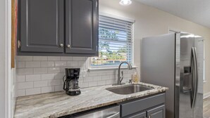 A kitchen with dark cabinets, a marble countertop, a stainless steel refrigerator, and a window with blinds. - Candlewood Casa ~ 3BR/2BA 15 Mins to Beach! (Navarre)