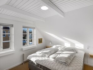 Ein Schlafzimmer mit schrägen Wänden, zwei Fenstern, einem Bett mit gesteppter Matratze und einem Nachttisch mit Lampe.