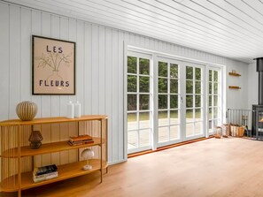 Ein Zimmer mit weißen Wandverkleidungen, einem großen Fenster mit Blick ins Grüne, einem Holzkonsolentisch mit Büchern und einer Vase sowie einem gerahmten Kunstwerk darüber.