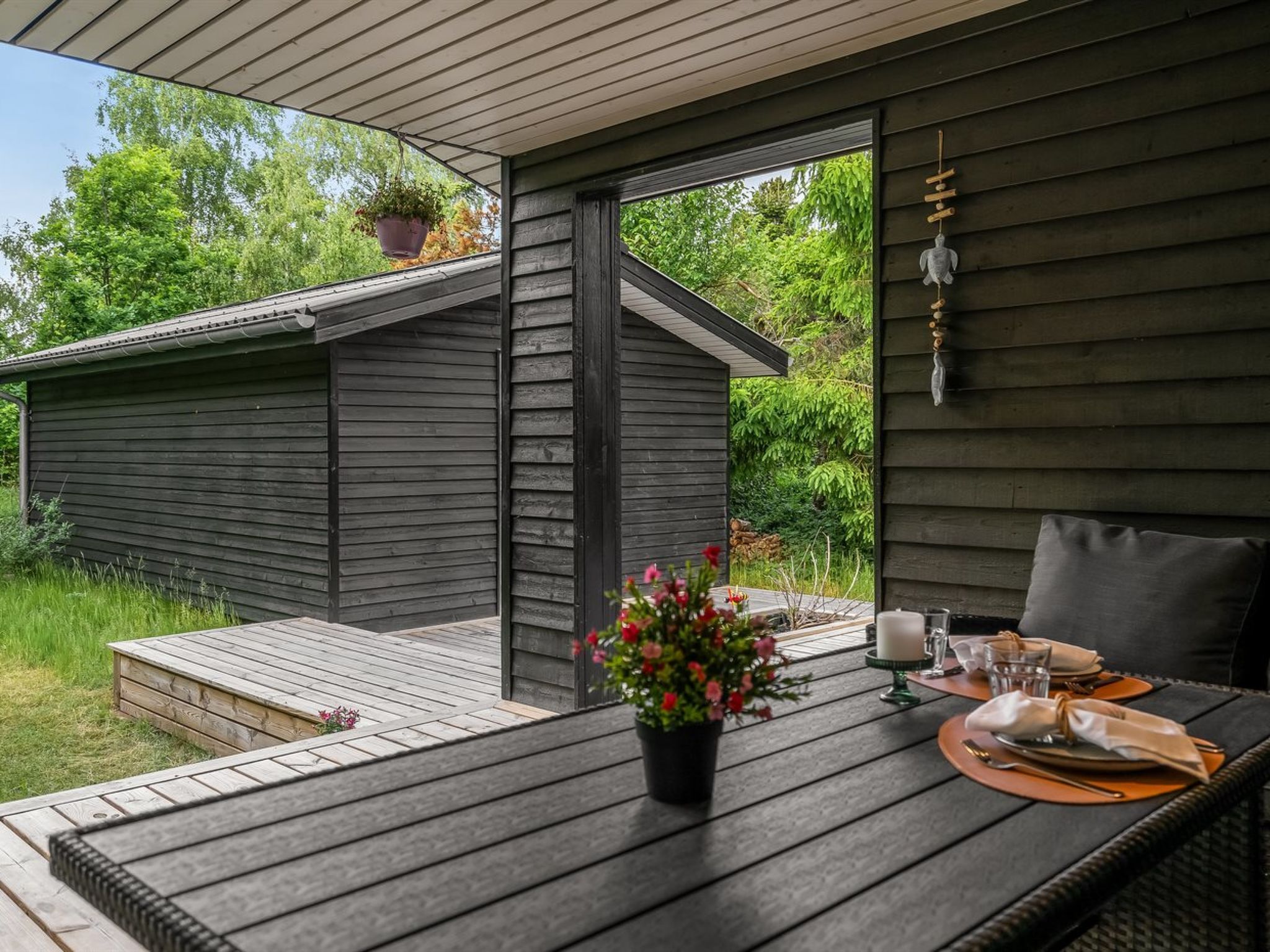 Eine Holzdar Terrasse mit einem Esstisch, ein kleines Gartenhaus im Hintergrund und ein Blumentopf auf dem Tisch.