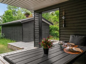Eine Holzdar Terrasse mit einem Esstisch, ein kleines Gartenhaus im Hintergrund und ein Blumentopf auf dem Tisch.
