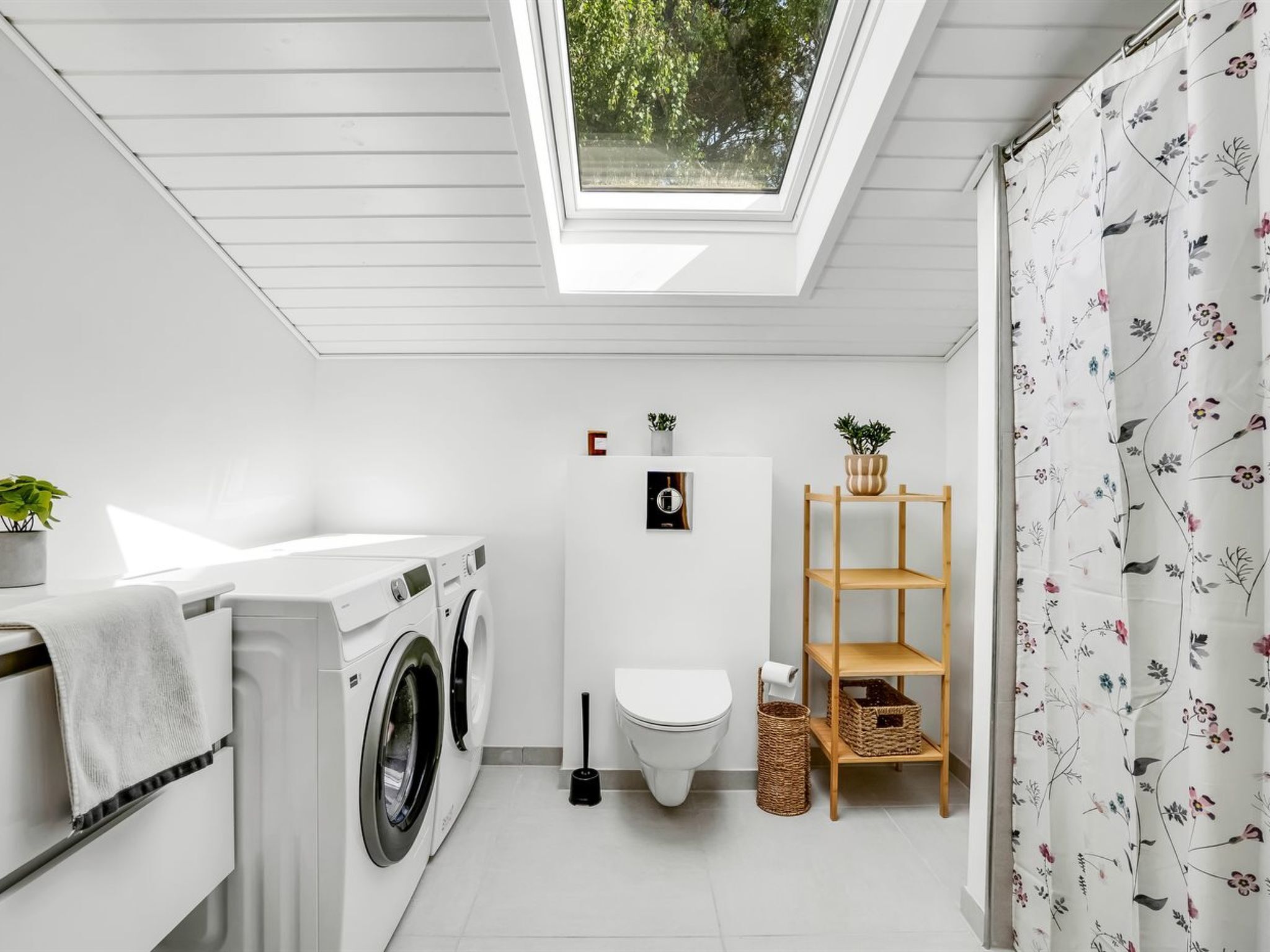 Een kleine wasruimte met dakraam, een wasmachine, toilet en een houten plank met planten.
