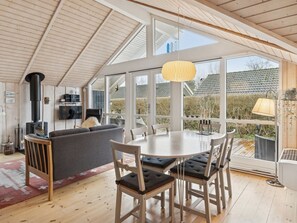 Een modern interieur met een hoge plafond, een open haard, een bank, een eettafel met stoelen en grote ramen met uitzicht op het omliggende landschap.