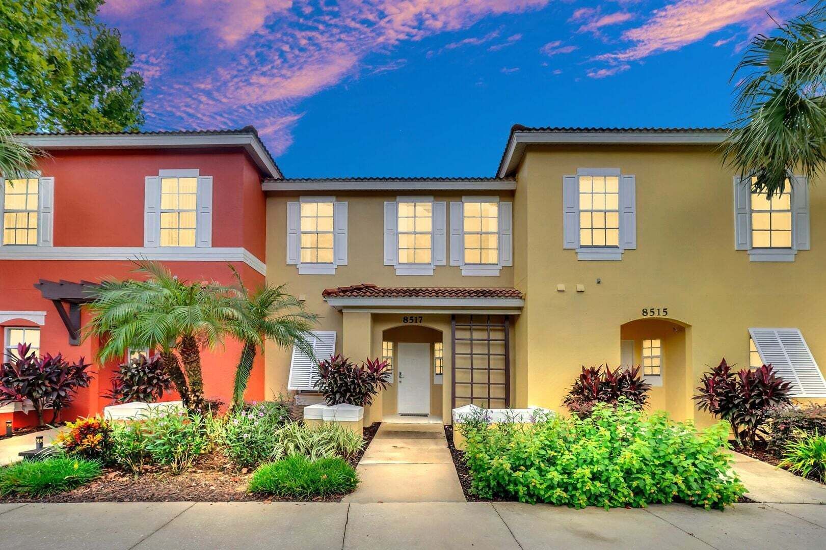 A row of three-story houses with a central entrance, numbered 8517, surrounded by well-maintained gardens and palm trees.