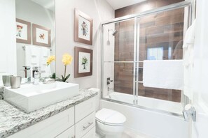 Une salle de bain moderne avec une douche à parois de verre, un lavabo blanc doté d’un plan de toilette en marbre et des œuvres d’art encadrées accrochées au mur.