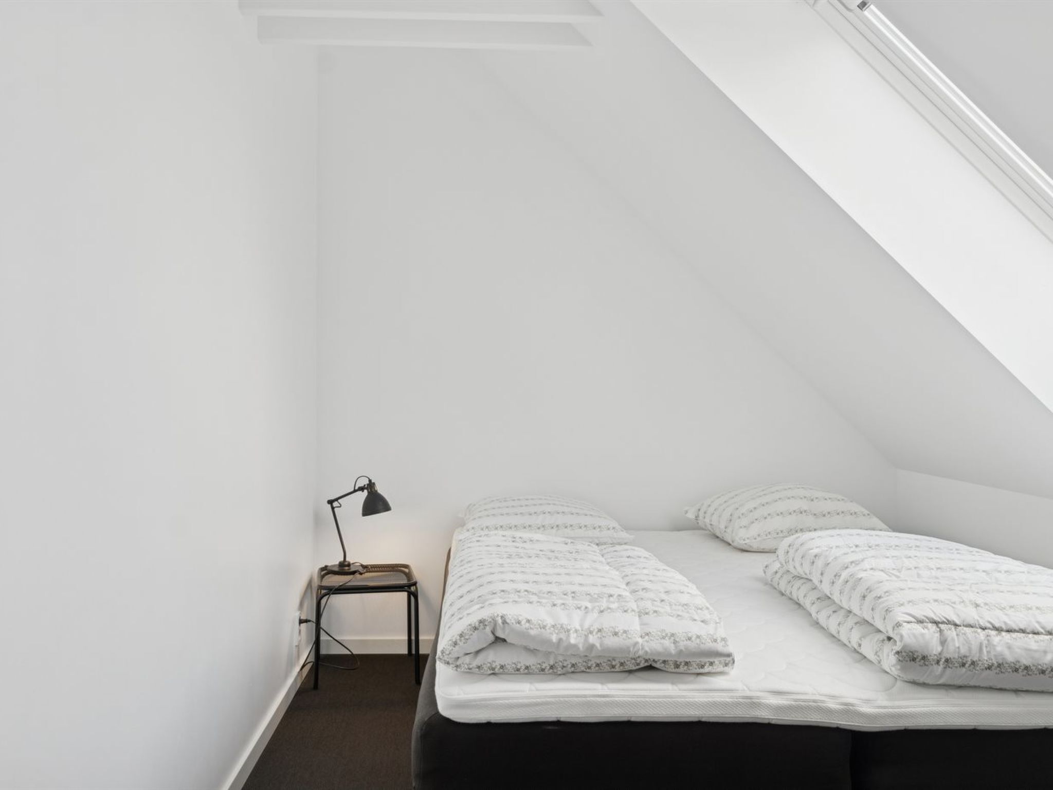 A small, single-bed room with a sloped ceiling, a nightstand with a lamp, and a window providing natural light.
