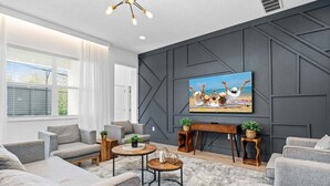 Un salon moderne avec des canapés gris, une table basse ronde, un téléviseur fixé à un mur foncé et une fenêtre avec des stores.