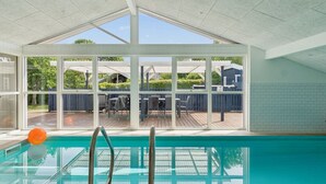 Een zwembad met glazen wanden, uitzicht op een terras met meubilair en een hoog plafond met dakramen.