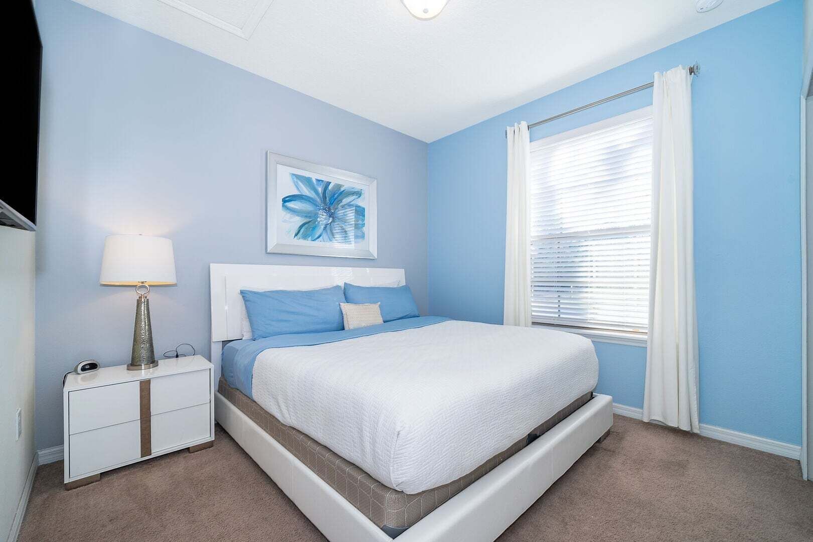 A bedroom with a bed, a nightstand, a lamp, a framed picture, and a window with curtains.